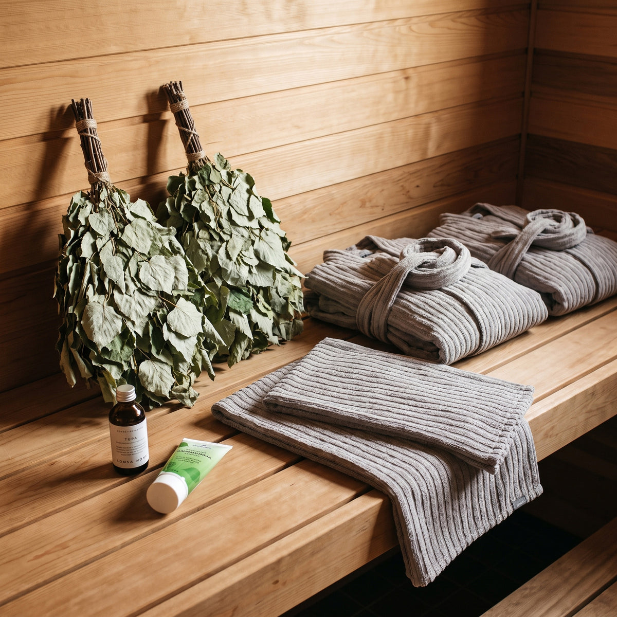 A cozy sauna scene with two leafy hand-woven brooms, folded gray ribbed bathrobes and towels, a small amber bottle, and a green-and-white tube on a wooden sauna bench against a wood plank wall.