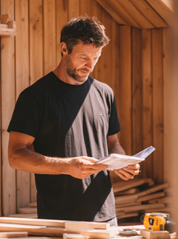 Man in a black t-shirt holding blueprints in a wooden workshop