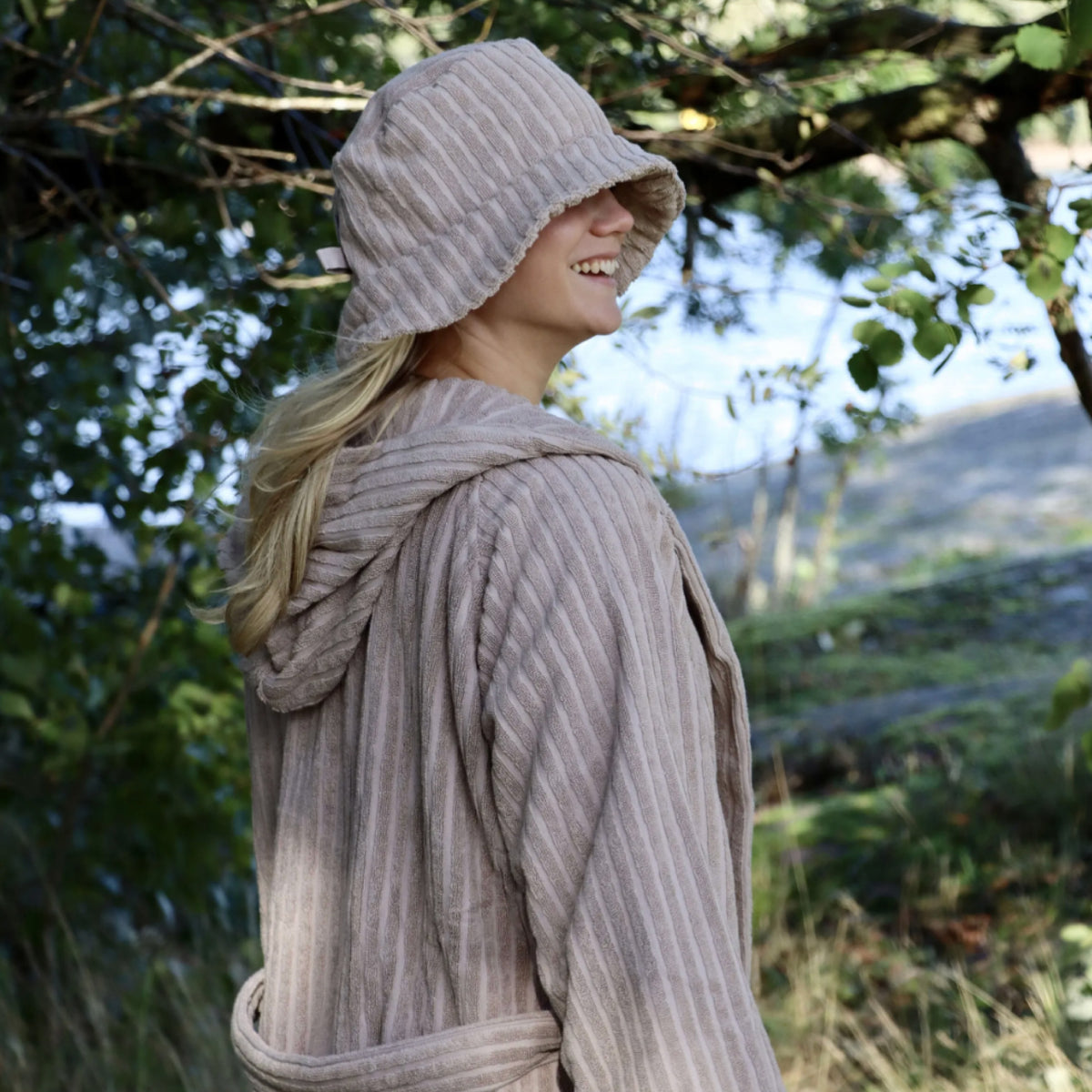 A woman outdoors wearing a matching ribbed knit hooded robe and bucket hat in a light taupe color, with trees and a blurred natural background.