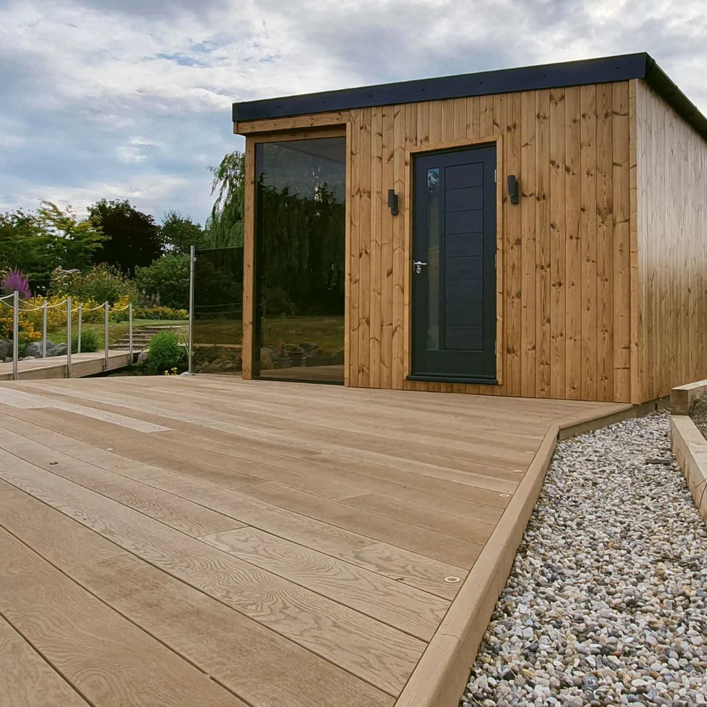 Wooden garden shed with a deck and gravel area, surrounded by greenery.