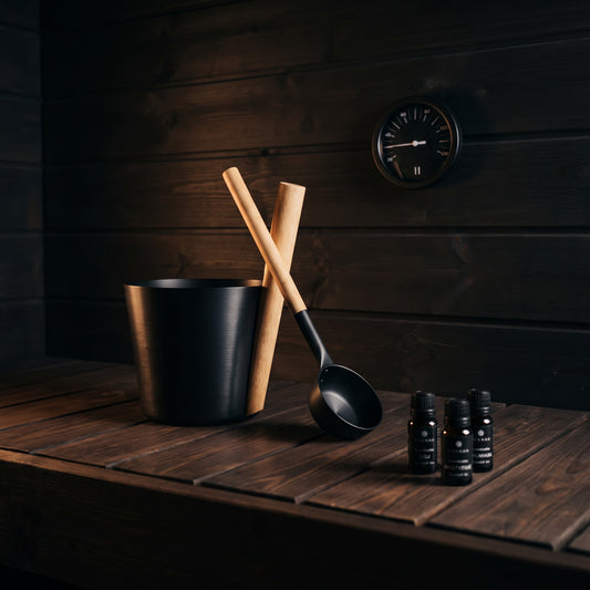 A black metal sauna bucket with two wooden-handled ladles resting inside, alongside three small black bottles, set on a dark wooden table against a dark wood-paneled wall.