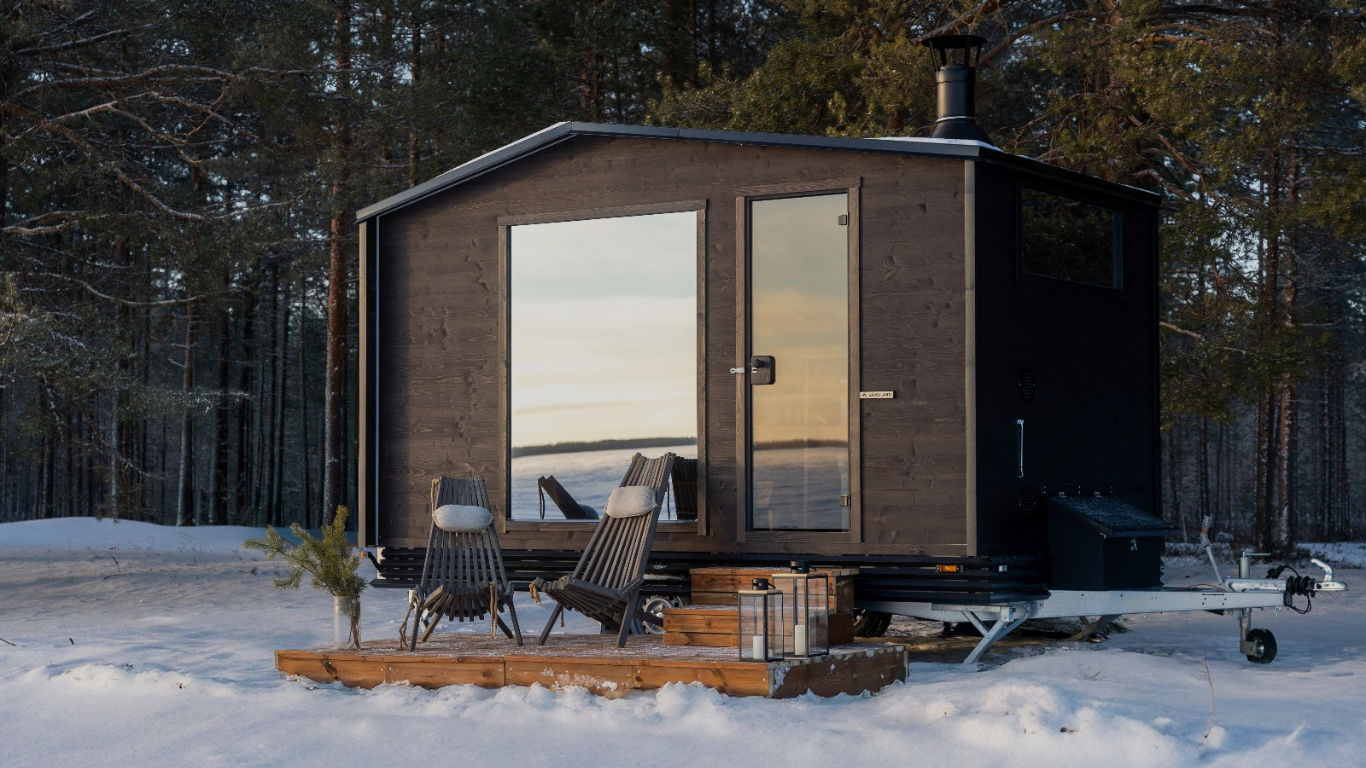 Small wooden cabin in a snowy forest with chairs and a table outside.