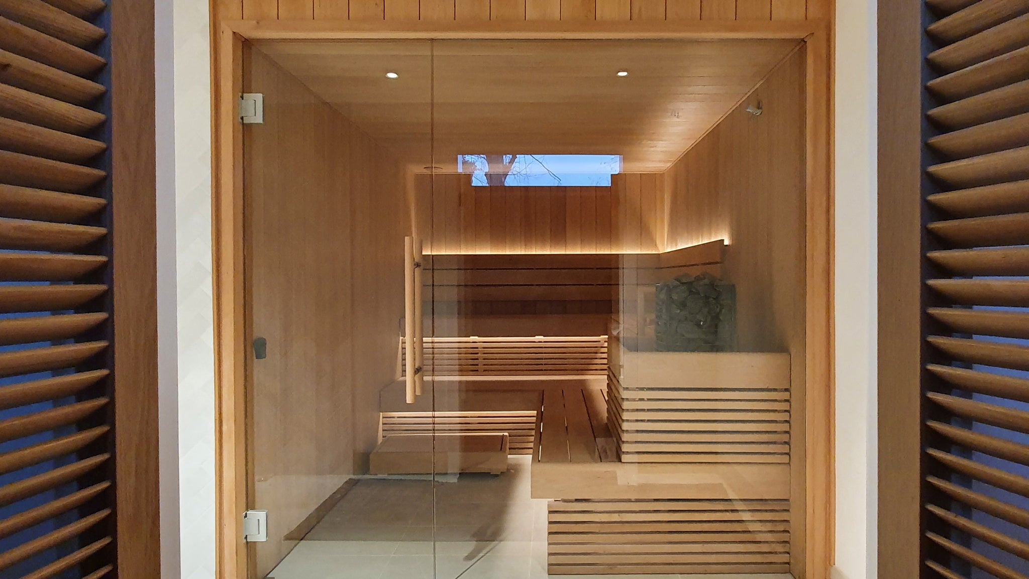 This Bespoke Alder Sauna Installation has a full glass wall, through which you can see a vertically clad sauna using alder tongue and groove cladding, an L shaped sauna bench system with an upper and lower bench and a Narvi Velvet Electric Sauna Heater.