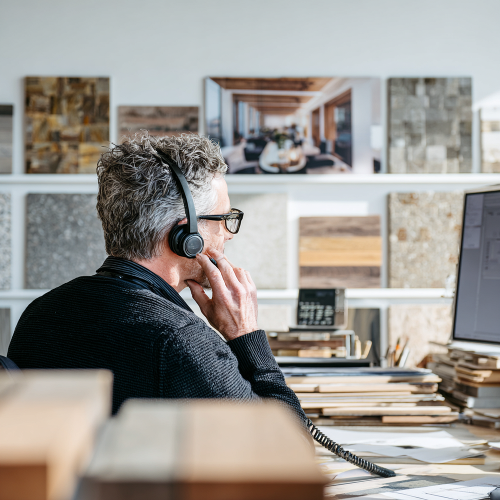 Man wearing headphones and talking on a phone in an office setting with wood samples in the background